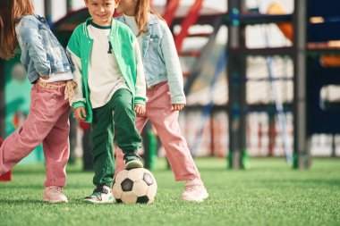 Yeşil elbiseli, futbol toplu bir çocuk. Çocuklar oyun alanında eğleniyor..