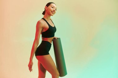 Siyah yoga minderi ellerinde. Spor elbiseli genç kadın stüdyoda arka planda karşı karşıya.