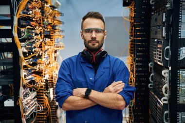 Kollarını kavuşturmuş duruyorsun. Genç adam sunucu odasında internet ekipmanı ve kablolarıyla çalışıyor..