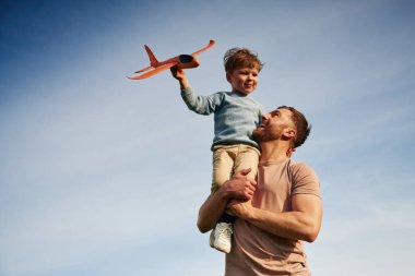 Beyaz bulutlu mavi gökyüzü. Baba oğlunu kucağında tutuyor, oyuncak uçakla oynuyor..