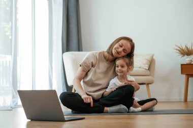 Laptop kullanıyorum. Küçük kızı olan genç bir kadın evde yoga yapıyor..