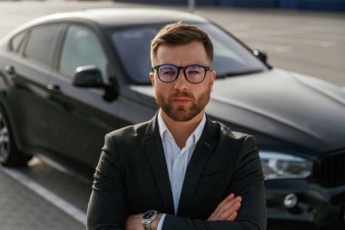 Gözlüklü, ön manzaralı. Takım elbiseli bir işadamı dışarıda siyah arabasının yanında..