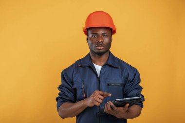 Dijital tablet elinde, inşaat işçisi. Yakışıklı siyah adam sarı arka plana karşı stüdyoda..