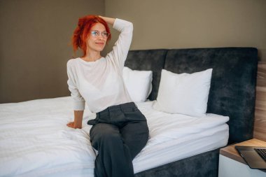 Resmi kıyafetlerle, yatakta oturarak. Kadın otel odasında, modern iç mekan..