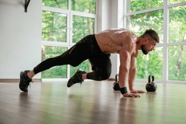 Pencerenin dışındaki yeşil ağaçlar. Güçlü kaslı adam spor salonunda çalışıyor..