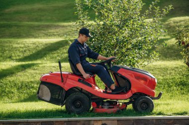 Kırmızı renkli çim biçme makinesi, üzerinde oturuyor. Bahçede profesyonel bahçıvan var..