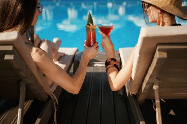 Çarpıcı bardaklar, lezzetli içecekler, dikiz aynası. İki güzel kadın havuz kenarında dinleniyor..