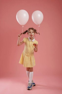 Dilini gösteriyor, şakacı, balonlarla. Şirin küçük kız pembe arka plana karşı..