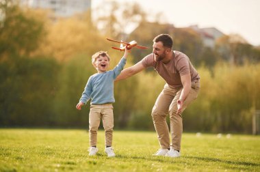 Günlük kıyafetlerle. Oyuncak uçakla koşuyor. Mutlu baba ve oğul yazın sahada eğleniyorlar..