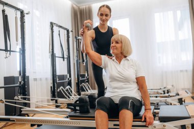 Dambıl nasıl kaldırılır. Kadın eğitmen yaşlı kadının egzersiz yapmasına yardım ediyor..