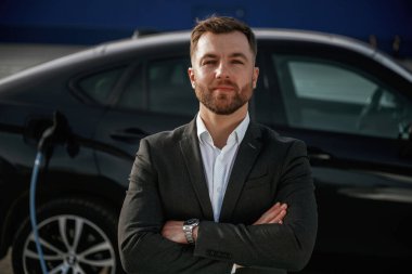 Kollarını kavuşturmuş duruyorsun. Takım elbiseli bir işadamı dışarıda siyah arabasının yanında..