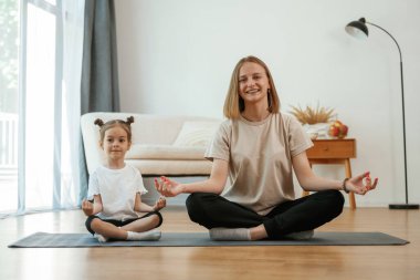 Gülümseyen, pozitif yüz ifadesi. Küçük kızı olan genç bir kadın evde yoga yapıyor..