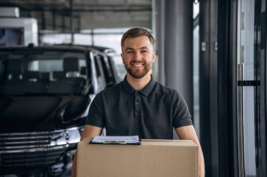 Gülümseyen, pozitif yüz ifadesi. Teslimatçı ya da yükleyici elinde bir kutuyla arabanın yanında..