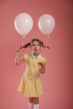 Dilini gösteriyor, şakacı, balonlarla. Şirin küçük kız pembe arka plana karşı..