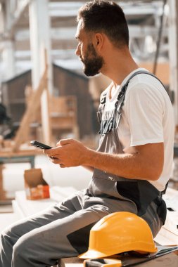 Dinleniyor, akıllı telefonla oturuyor. Ahşap depoda sanayi işçisi..