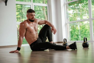 Pencerenin dışındaki yeşil ağaçlar. Güçlü kaslı adam spor salonunda çalışıyor..