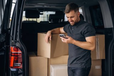 Akıllı telefonu kullanıyordu. Teslimatçı ya da yükleyici elinde bir kutuyla arabanın yanında..