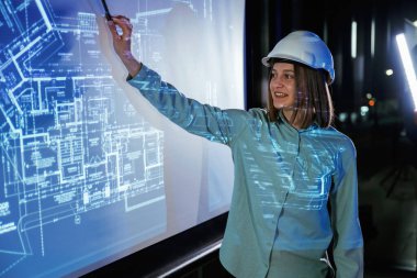 Beyaz kasketli. Kadın mimar projektör üzerine inşaat alanı planı gösteriyor.