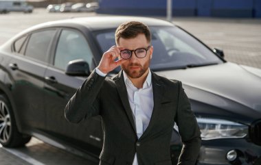 Gözlüklü, ön manzaralı. Takım elbiseli bir işadamı dışarıda siyah arabasının yanında..