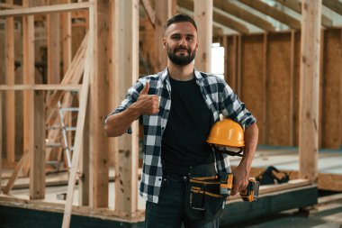 Evi inşa ediyor, elinde şapka ve matkapla. Ahşap depoda sanayi işçisi..