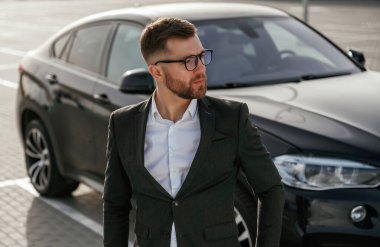 Gözlüklü, ön manzaralı. Takım elbiseli bir işadamı dışarıda siyah arabasının yanında..