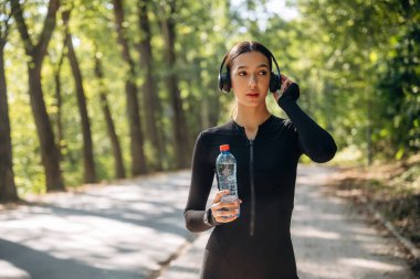 Mola verdim. Kulaklıklar ve suyla ayakta durmak. Genç, sıska kadın dışarıda siyah spor kıyafetleri giyiyor..