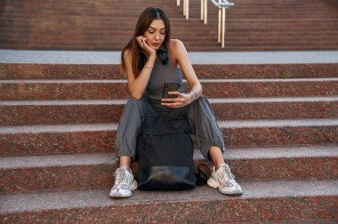 Akıllı telefonu tutarak, merdivenlerde oturarak. Güzel kız öğrenci üniversitenin yakınında dışarıda..