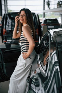 Aracın üzerinde duruyor ve yaslanıyor. Güzel genç bayan araba galerisinde yeni arabasıyla..