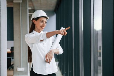 Parmaklarıyla kenarı işaret ediyor. Kasketli ve beyaz elbiseli kadın mimar içeride..