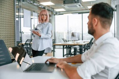 Patron dizüstü bilgisayarla masada oturuyor. Kadın ve erkek ofiste birlikte çalışıyorlar..