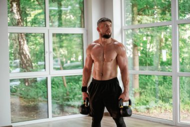 Kalkıyor, dambılları tutuyor. Güçlü kaslı adam spor salonunda çalışıyor..