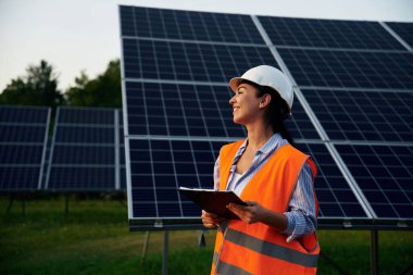 Elinde siyah bir not defteri var. Üniformalı kadın işçi mühendisi güneş panellerinin yakınında..