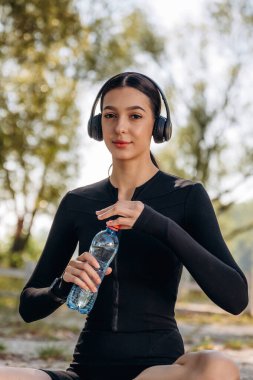 Rahatlamış, yoga minderinde oturuyor, mola. Genç, sıska kadın dışarıda siyah spor kıyafetleri giyiyor..