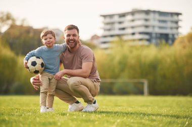 Oturuyor, futbol topu tutuyor. Mutlu baba ve oğul yazın sahada eğleniyorlar..