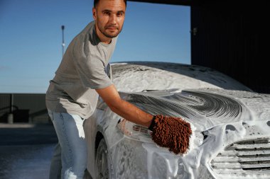 Otomotiv temizleyicisi sabun ve köpüğü yüksek basınçlı çamaşır makinesiyle yıkıyor. Adam dışarıda arabasıyla bekliyor..