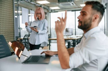 Patron dizüstü bilgisayarla masada oturuyor. Kadın ve erkek ofiste birlikte çalışıyorlar..