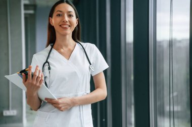 Ayakta duran ve belgeleri elinde tutan. Beyaz önlüklü kadın doktor içeride..