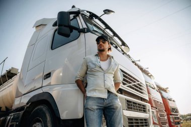 Aşağıdan bak, ayakta ve poz veriyor. Kamyon şoförü açık havada nakliye aracının yanında..