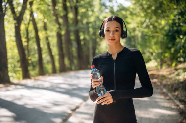 Mola verdim. Kulaklıklar ve suyla ayakta durmak. Genç, sıska kadın dışarıda siyah spor kıyafetleri giyiyor..