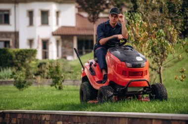 Çim biçme makinesinin üzerinde oturuyor. Bahçede profesyonel bahçıvan var..