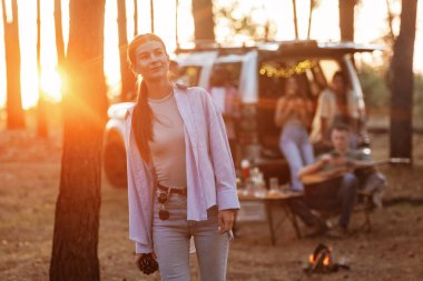 Genç bayan ayakta duruyor ve poz veriyor. Bir grup arkadaş ormanda birlikte eğleniyorlar..