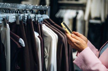 Akıllı telefondan bilgi okuyorum. Perakende dükkanında kıyafet seçen bir kadının yakın görüntüsü..