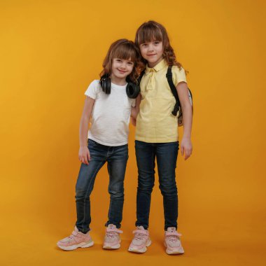 İki çocuk arkadaş ayakta, birbirlerine sarılıyorlar ve sarı arka planda poz veriyorlar..