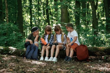 Dinleniyor, kamera ve su şişesini tutuyor. Çocuklar yazın gündüz vakti ormanda..