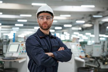 Kollarını kavuşturmuş poz veriyor. Fabrika işçisi kasketli bir şekilde içeride..