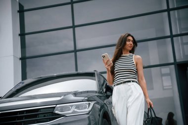 Akıllı telefonlu. Çekici genç bayan dışarıda, yepyeni arabasının yanında..