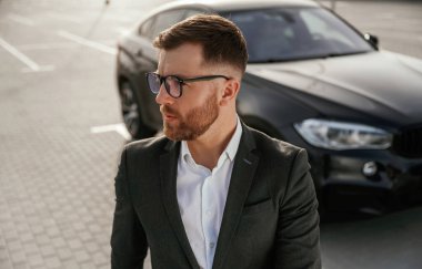 Gözlüklü yakışıklı, zeki adam. Takım elbiseli bir işadamı dışarıda siyah arabasının yanında..