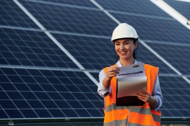 Not defteriyle verileri kontrol ediyorum. Üniformalı kadın işçi mühendisi güneş panellerinin yakınında..
