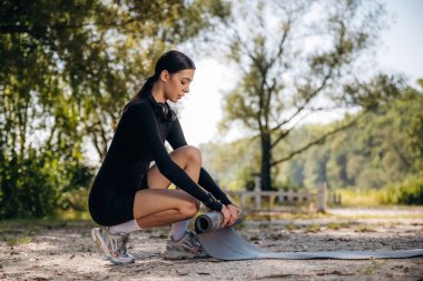 Yoga minderinden yere yuvarlanıyor. Genç, sıska kadın dışarıda siyah spor kıyafetleri giyiyor..