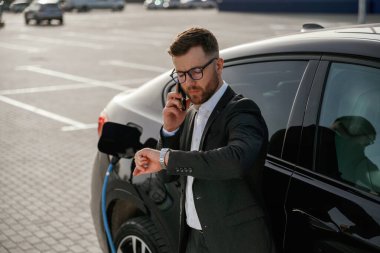 Şarj olurken araca yaslan, akıllı telefonla. Takım elbiseli bir işadamı dışarıda siyah arabasının yanında..